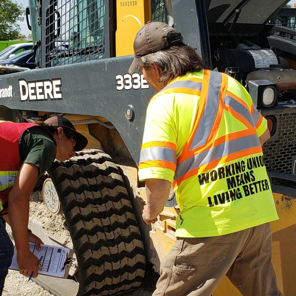 Two workers in high-visibility vests carrying out a pre-use inspection on a John Deere 333E tracked skid steer, one filling in an inspection checklist, during skid steer train-the-trainer in Manitoba