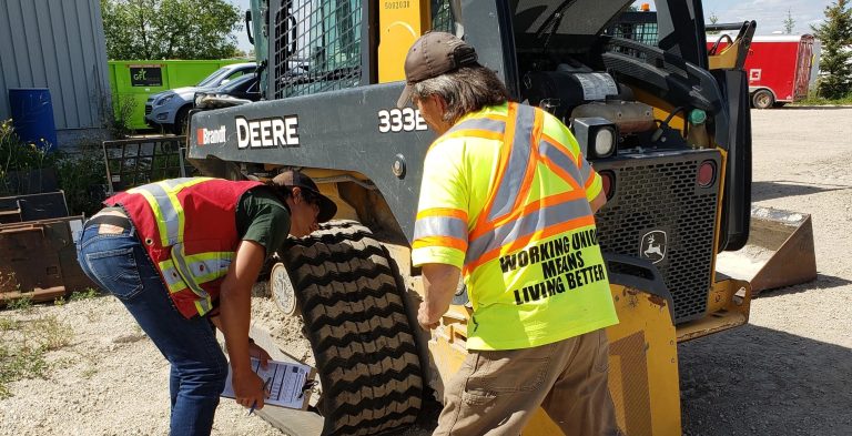 Two workers in high-visibility vests carrying out a pre-use inspection on a John Deere 333E tracked skid steer, one filling in an inspection checklist, during skid steer train-the-trainer in Manitoba