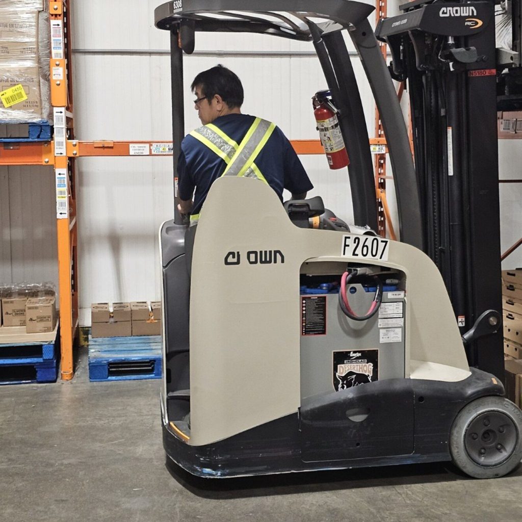 Operator in a hi-vis vest driving a stand-up reach truck down a warehouse aisle during on-site training in Winnipeg